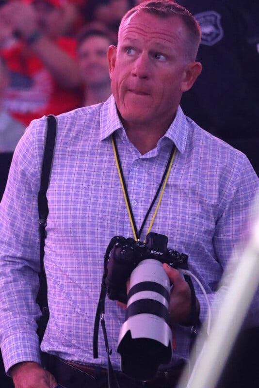 A man in a checkered shirt with a camera and long lens around his neck looks upward in a crowded indoor setting, wearing a yellow lanyard and shoulder strap.