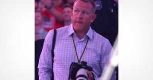 A man in a checkered shirt with a camera slung across his body stands in a crowd, looking slightly upward with a thoughtful expression. The background is blurred, showing people and an officer.
