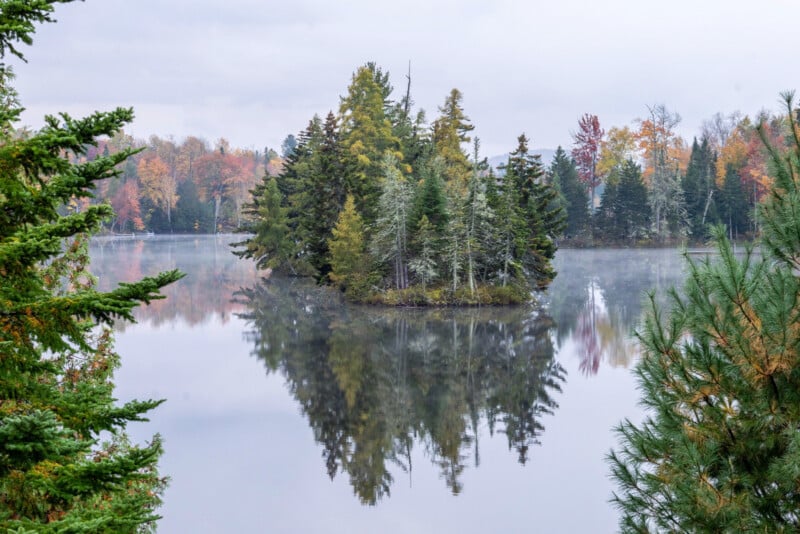A small forested island with evergreen trees is reflected in a calm lake, surrounded by autumn-colored trees and light mist in the background.