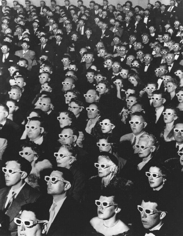 A large audience sits in rows, all wearing 3D glasses and facing forward, watching something off-frame in a black and white photograph.