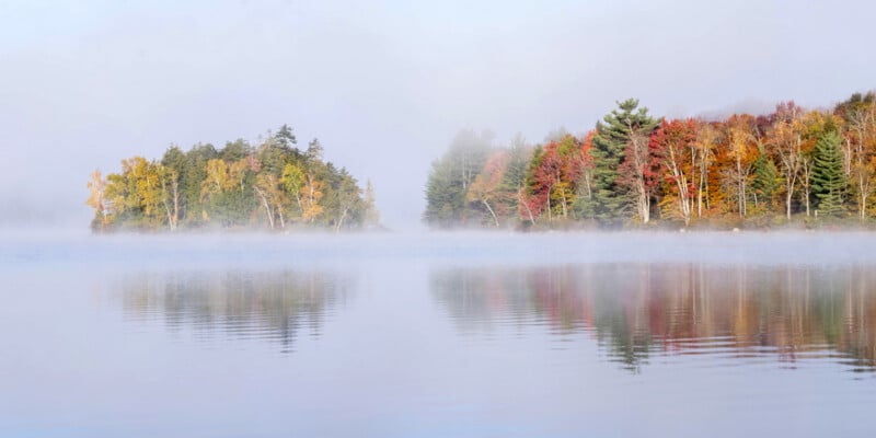 A calm lake reflects a misty scene of autumn trees with vibrant red, yellow, and green foliage on the shoreline. The fog creates a soft, tranquil atmosphere and blurs the background.