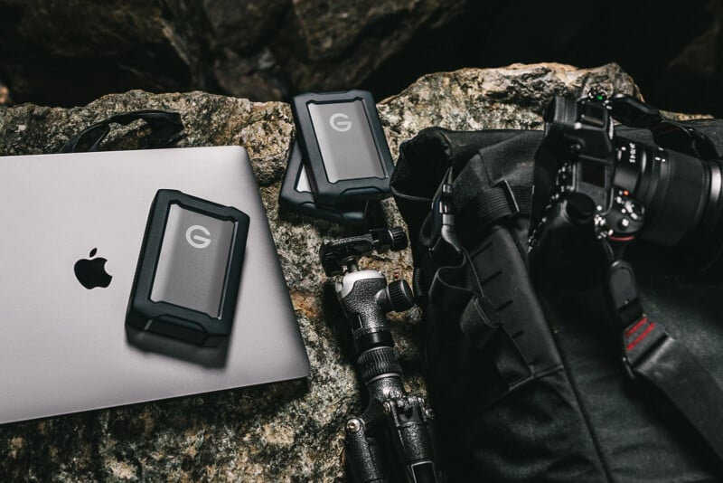 A MacBook, two rugged external hard drives, a camera, a camera lens, and a black backpack are placed on large rocks, suggesting an outdoor photography setup or workspace.