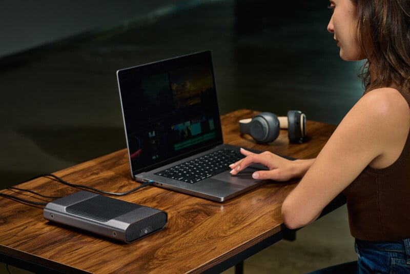 A woman sits at a wooden table using a laptop with a large external device connected. Wireless headphones and a smartwatch are also on the table. The workspace is modern and minimalistic.