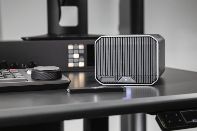 A compact, rectangular black device with vertical ridges sits on a desk beside a keyboard, a large control knob, and a pen. Background shows buttons and blurred office equipment.