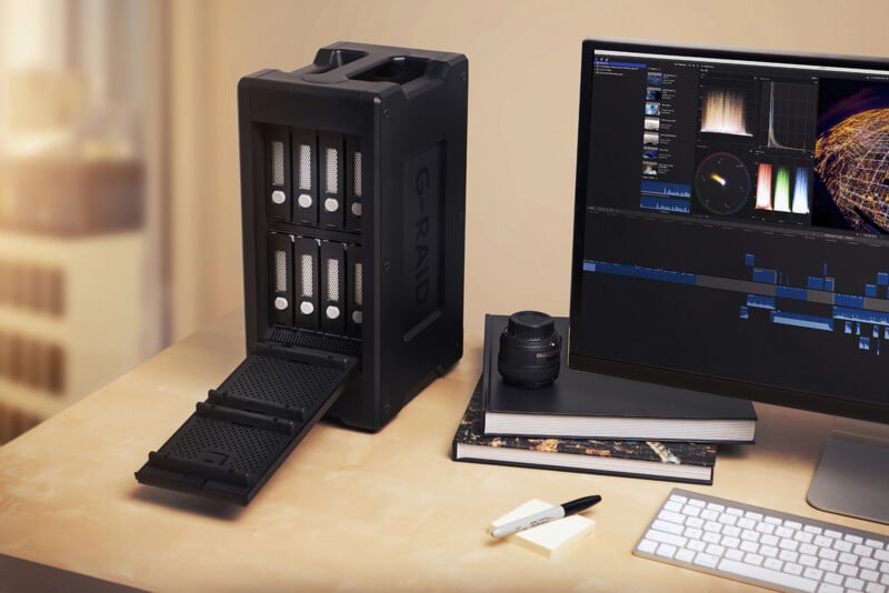 A desktop with a large external hard drive enclosure with open slots, a computer monitor displaying video editing software, a camera lens, stacked books, a pen, and note paper. Sunlight streams in from the left.