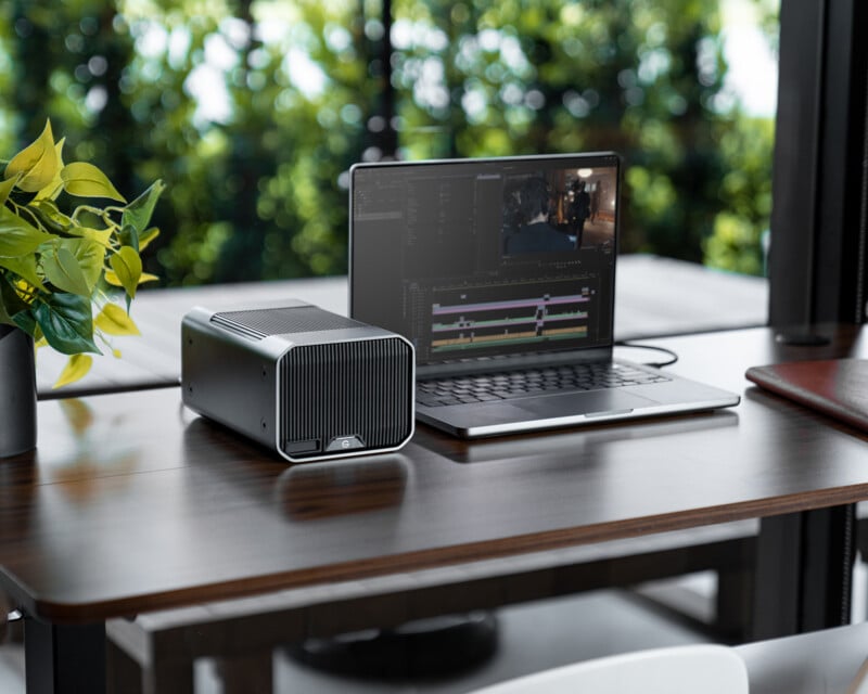 A laptop with video editing software open is on a wooden desk next to a compact desktop computer. A plant and a closed notebook are also on the desk, with greenery visible outside the window in the background.