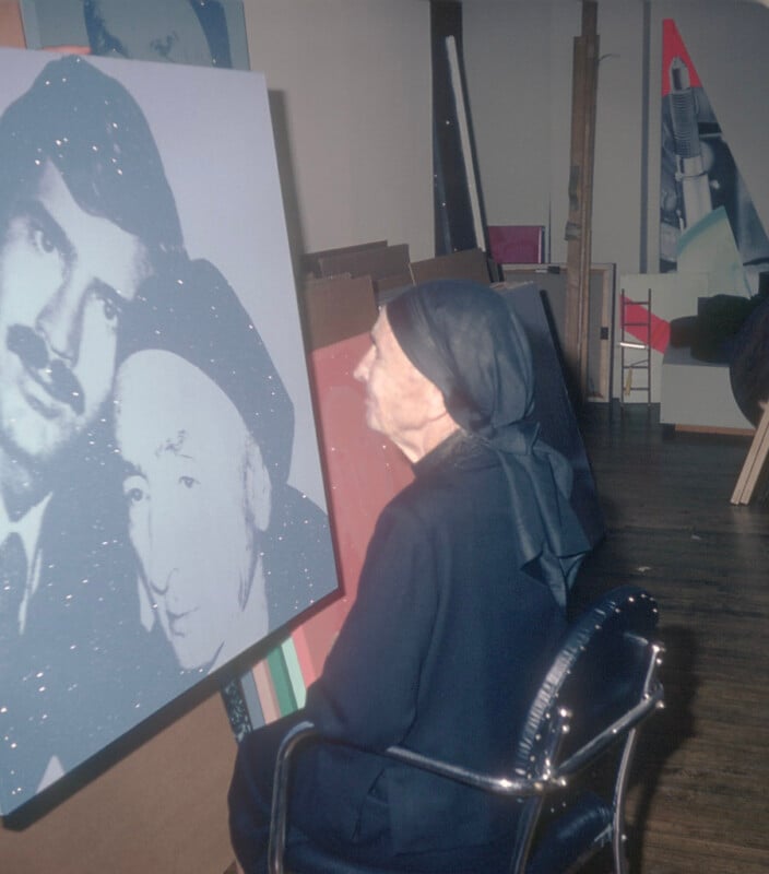 An elderly person wearing a dark headscarf sits in a chair, looking closely at a large canvas with two black-and-white portraits in an art studio filled with paintings and art supplies.