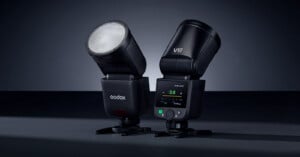 Two black Godox V1 camera flashes are positioned side by side on a dark surface. One shows its illuminated round head, while the other displays its digital control screen and buttons.