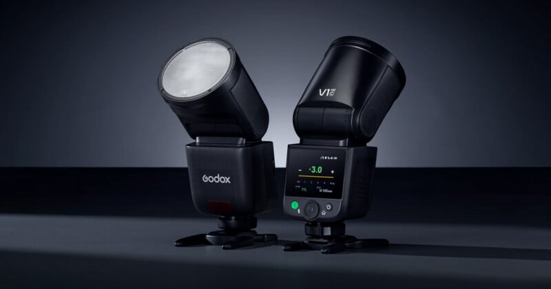 Two black Godox V1 camera flashes are positioned side by side on a dark surface. One shows its illuminated round head, while the other displays its digital control screen and buttons.