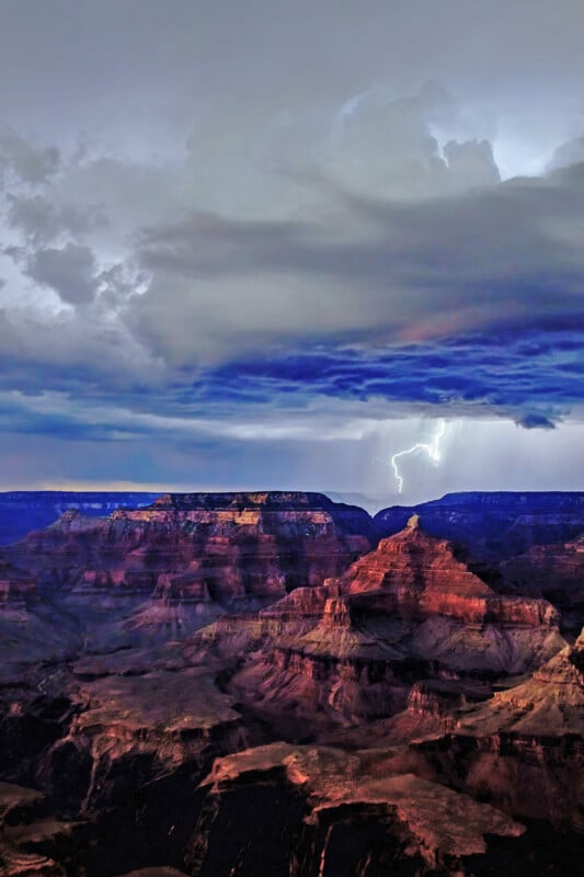 Dark storm clouds gather above the Grand Canyon as a bright bolt of lightning strikes in the distance, illuminating the dramatic rock formations and layered cliffs below.