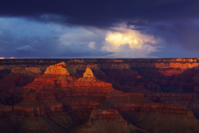 Sunlight illuminates the layered red rock formations of the Grand Canyon under a dramatic sky, with dark storm clouds and a patch of bright light breaking through in the distance.