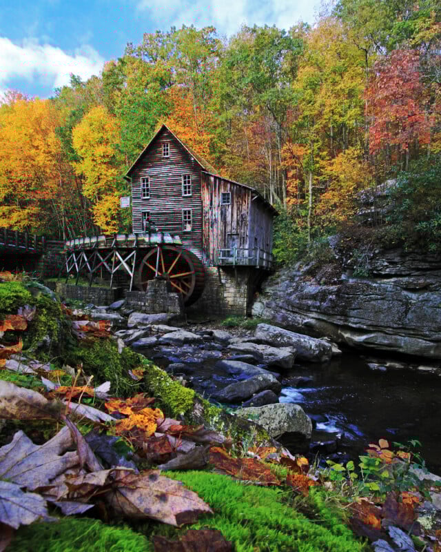 A rustic wooden watermill with a large water wheel stands by a rocky stream, surrounded by vibrant autumn trees with colorful foliage and mossy rocks in the foreground.