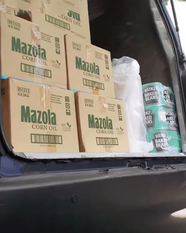 Stacked boxes of Mazola corn oil and packs of Heinz Baked Beans inside the open back of a dark vehicle, with additional plastic-wrapped goods visible on the right side.