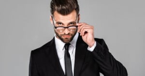 A man in a black suit and tie looks seriously at the camera while adjusting his glasses with one hand, against a plain gray background.