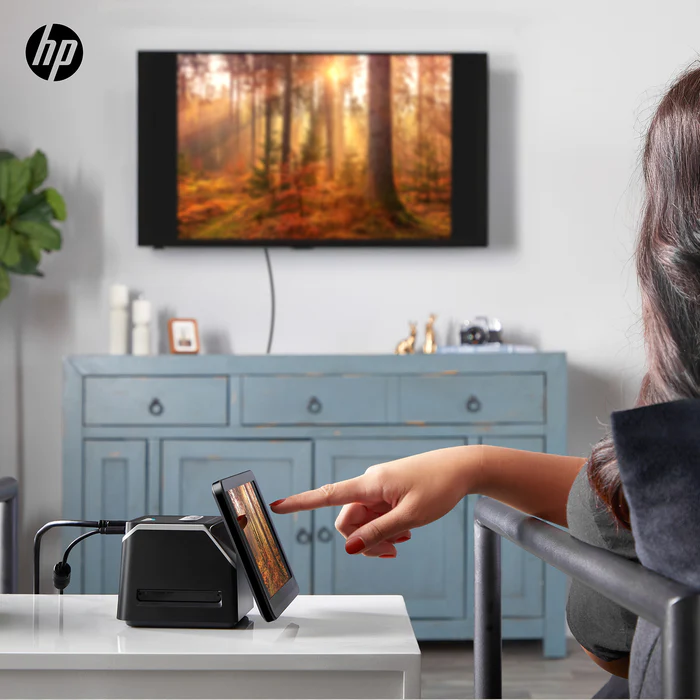 Una persona se sienta en una sala de estar y utiliza un dispositivo de pantalla táctil colocado sobre la mesa. El dispositivo se conecta mediante un cable y refleja su pantalla en un gran televisor montado en la pared que muestra una escena del bosque. El logotipo de HP aparece en la esquina.
