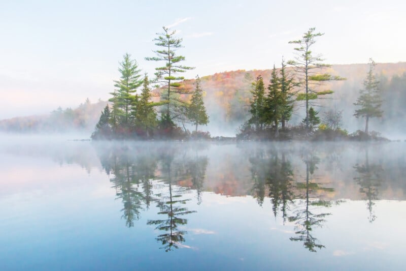 A small cluster of evergreen trees stands on a misty lakeshore, their reflections mirrored in the calm, glassy water. Soft morning light and gentle fog create a peaceful, serene atmosphere with distant hills in the background.