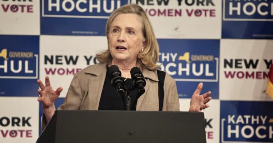 A woman speaks at a podium with microphones, gesturing with her hands. Behind her are campaign signs that read "Kathy Hochul," "New York Governor," and "Women Vote.