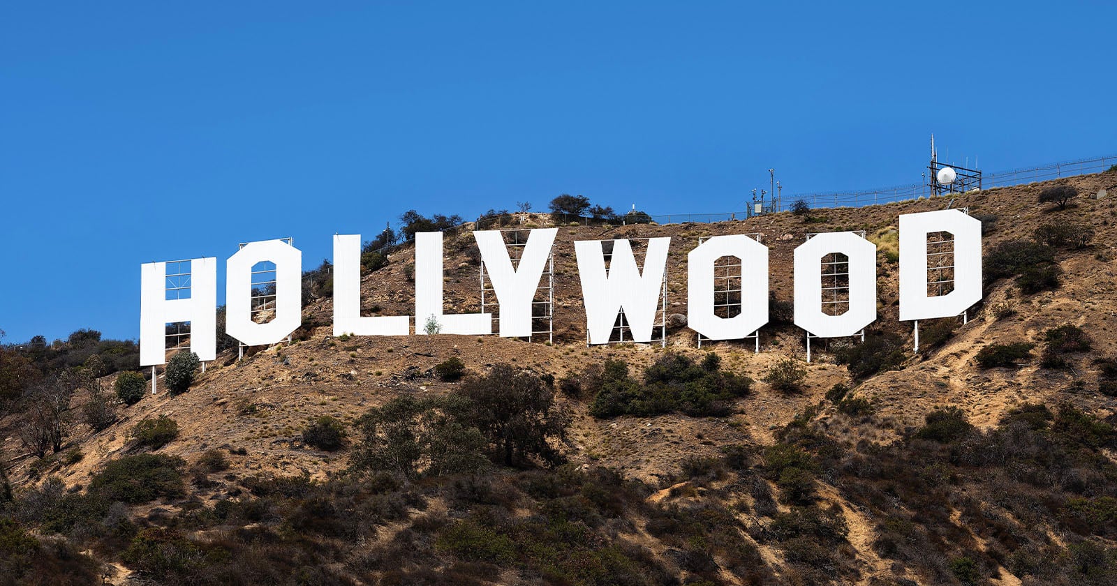 Large white letters spelling "HOLLYWOOD" stand on a dry, shrub-covered hillside under a clear blue sky. The famous landmark is set against rugged terrain with sparse vegetation.
