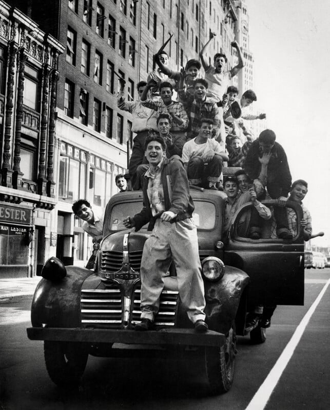 A group of excited young men pose and cheer on top of and around an old pickup truck on a city street, with tall buildings in the background.