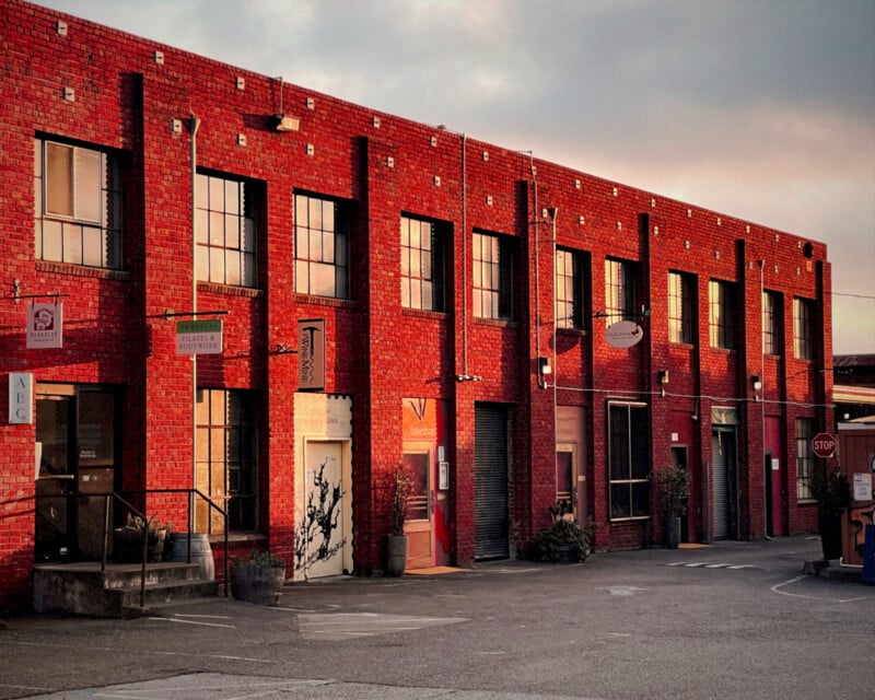 Una hilera de edificios industriales de ladrillo rojo de dos pisos con grandes ventanales y varios carteles comerciales proyectan una luz cálida y largas sombras en las paredes exteriores bajo el sol poniente. Hay plantas en macetas y una señal de alto enfrente.