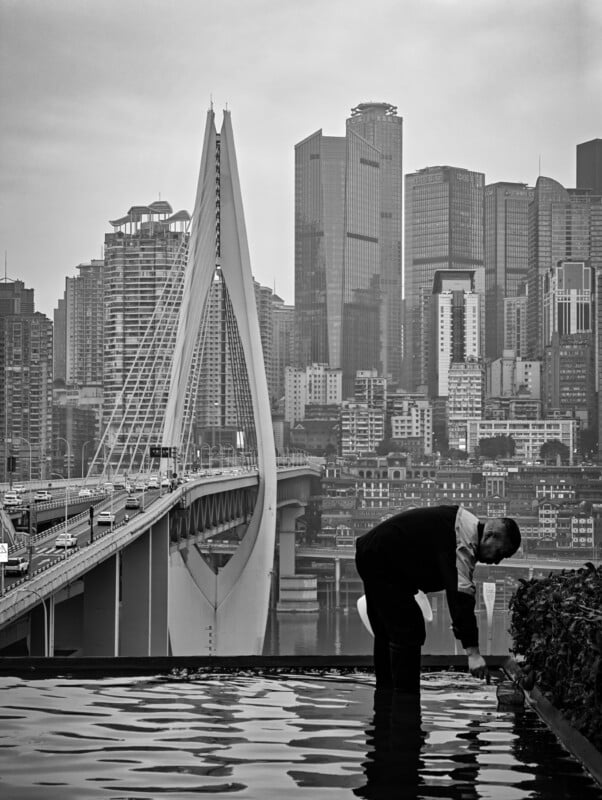 A person bends over a reflective pool in the foreground, with a modern suspension bridge and tall city skyscrapers in the background under a cloudy sky. The image is in black and white.