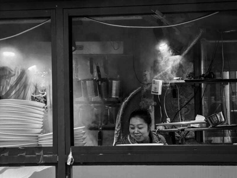 A woman stands behind the counter of a busy kitchen, surrounded by stacks of plates, hanging utensils, and steam rising into the air. The scene is captured in black and white.