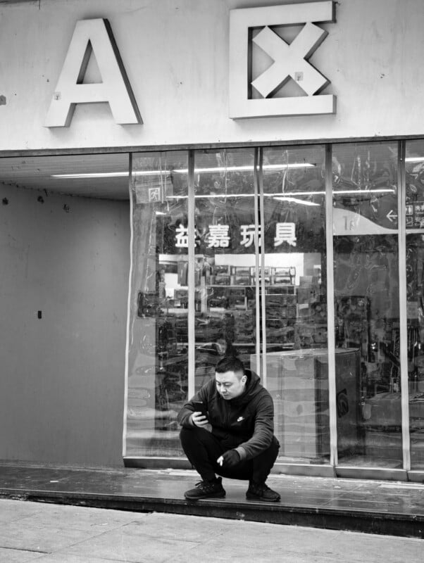 A man in dark clothing squats on a sidewalk, looking at his phone in front of a shop with large signs and glass windows reflecting shelves inside. The image is in black and white.