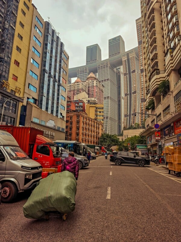 A person pulls a large green-wrapped bundle on a cart along a busy street lined with tall buildings and parked vehicles, with modern skyscrapers visible in the background under an overcast sky.