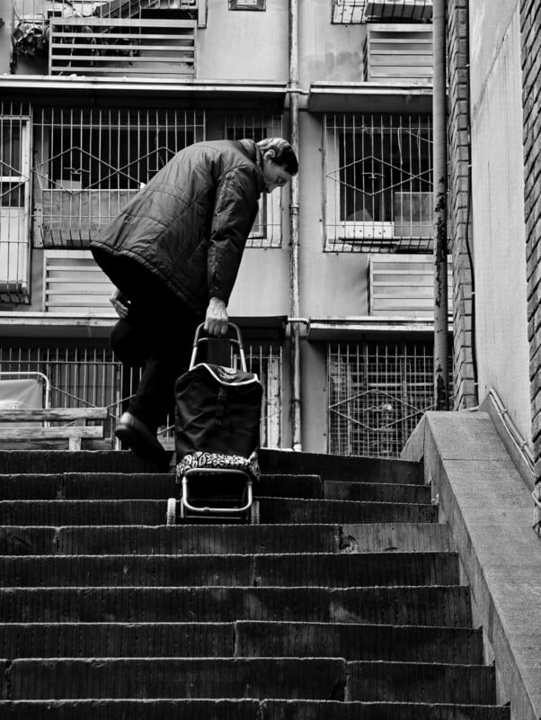An elderly person in a coat pulls a shopping trolley up a set of outdoor stairs in front of an apartment building with barred windows. The image is in black and white.