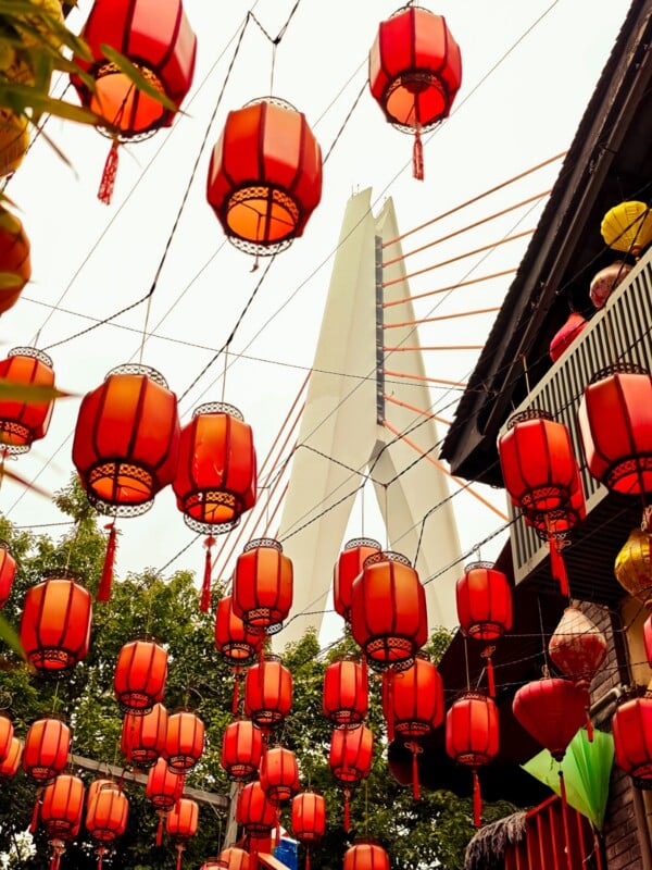 Red Chinese lanterns hang overhead with a modern, pointed white monument in the background. Trees and a building with balconies are also visible, creating a festive outdoor scene.