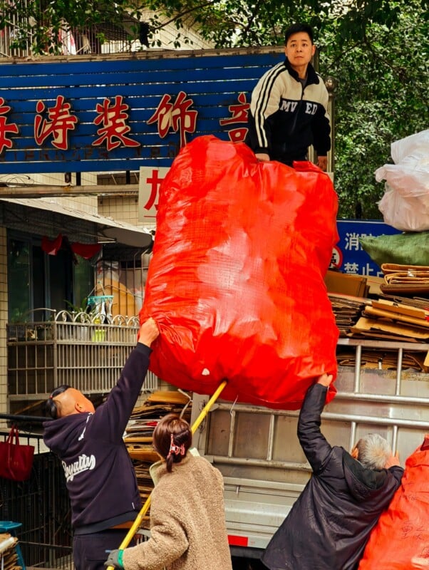 Three people on the ground lift a large red sack onto a truck, while a man on the truck guides it. They are in an urban area with signs and buildings in the background.