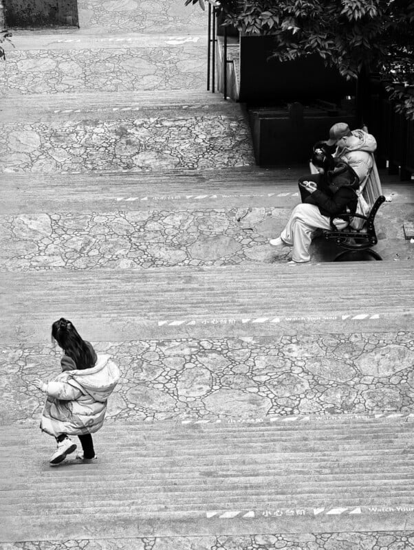 Black and white photo of wide stone steps; a child walks alone at the bottom left, while two people sit on a bench to the right, partially under a tree. The steps have cracks and faint text reading "Watch Your Step.