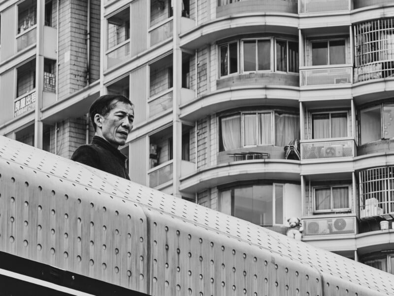 A man stands behind a perforated barrier, looking to the side, with a high-rise apartment building featuring numerous windows and balconies in the background. The image is in black and white.