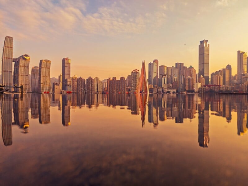 A modern city skyline at sunset is reflected on calm water, with tall skyscrapers, high-rise buildings, and an orange sky creating a warm, tranquil urban scene.