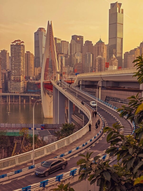 A curving bridge leads into a modern city with tall skyscrapers at sunset. Cars and pedestrians travel along the bridge, with a river and more buildings visible in the background. Trees partially frame the scene.