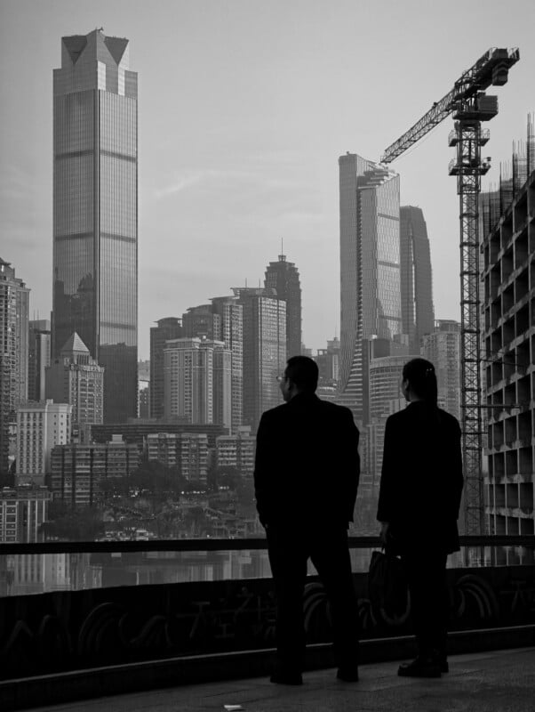 Two people in business attire stand on a balcony, looking at a city skyline with tall skyscrapers, modern buildings, and a construction crane, all in black and white.