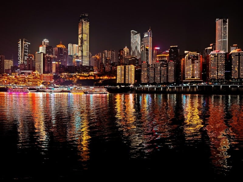 A vibrant city skyline at night with tall illuminated buildings and colorful lights reflecting on the calm river below.