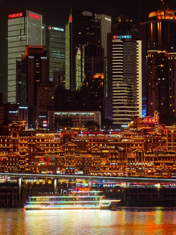 A brightly lit cityscape at night features a glowing river cruise boat on the water, rows of illuminated traditional-style buildings, and tall modern skyscrapers in the background.
