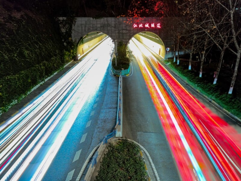 Long-exposure photo of a road tunnel at night, showing bright white and red light trails from cars passing through, with illuminated Chinese characters above the tunnel entrances. Trees and greenery line the roadside.
