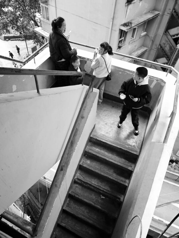 Four people stand on an outdoor concrete staircase landing; two women face each other while looking at their phones, and two children stand nearby. Buildings and street scenes are visible in the background.