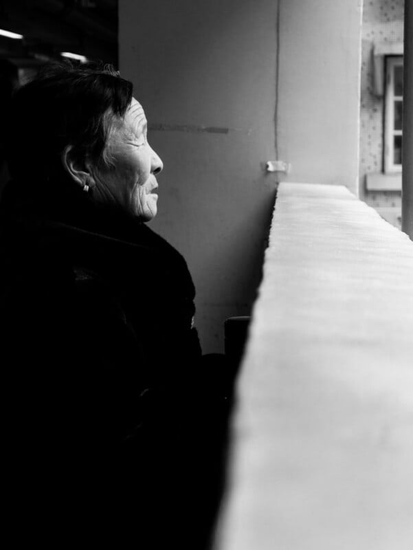 An elderly person with short hair and earrings gazes out of a window in profile. The black-and-white photo emphasizes light on their face while the foreground and background remain softly out of focus.