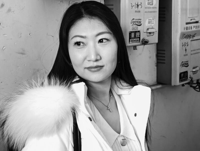 A woman with long dark hair, wearing a white coat with a fur collar, stands in front of a wall with meters and signs. She is looking slightly to the side with a gentle expression. The photo is in black and white.