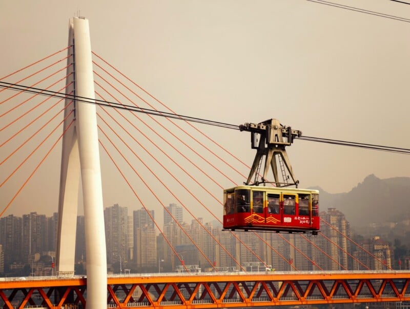 A red cable car travels on wires above a cityscape with tall buildings, a white cable-stayed bridge with orange cables, and hazy mountains in the background.