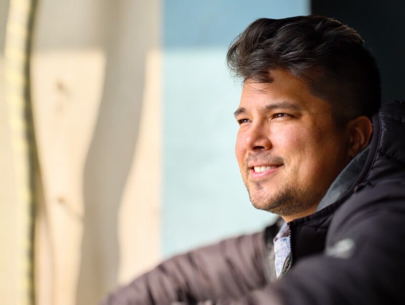 A man with short, dark hair and a slight beard smiles while looking out of a window. He is wearing a dark jacket and appears relaxed, with soft natural light illuminating his face.