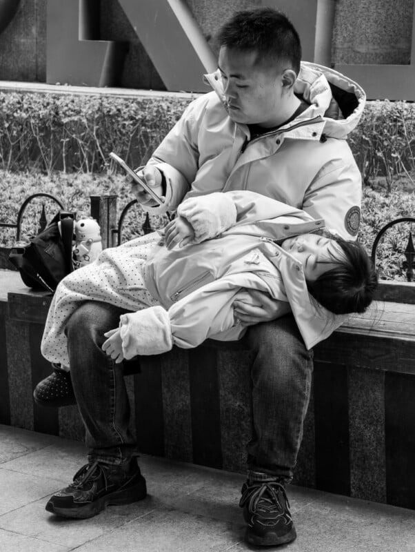 A man in a jacket sits on a bench, holding a sleeping child in his lap while looking at his phone. The child is bundled in a coat, lying comfortably with eyes closed. Bags sit beside them. The scene is in black and white.