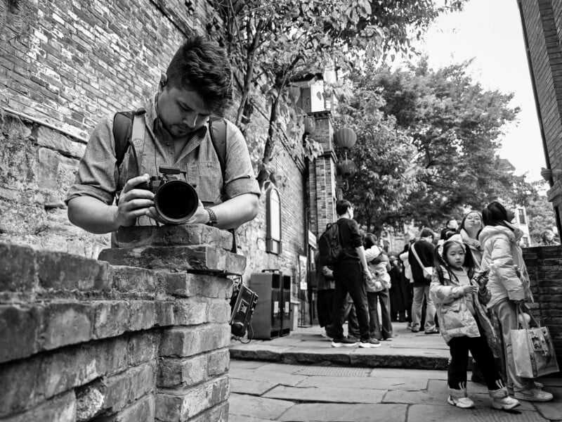A man with a camera adjusts his gear on a brick wall in a bustling street as a crowd, including children and adults, stands in the background. The scene is lively and set in an old, tree-lined alleyway.