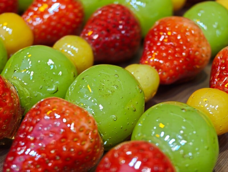 Close-up of glossy, colorful fruit skewers featuring strawberries, green grapes, and yellow fruit, all coated in a shiny, translucent glaze.