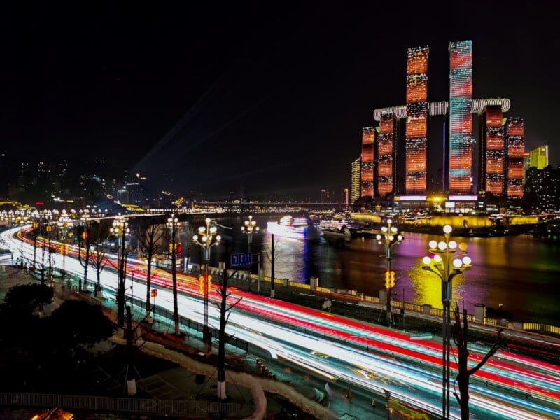 A vibrant cityscape at night with tall, illuminated buildings by a river and bright light trails from moving vehicles on a busy road, reflecting urban energy and motion.