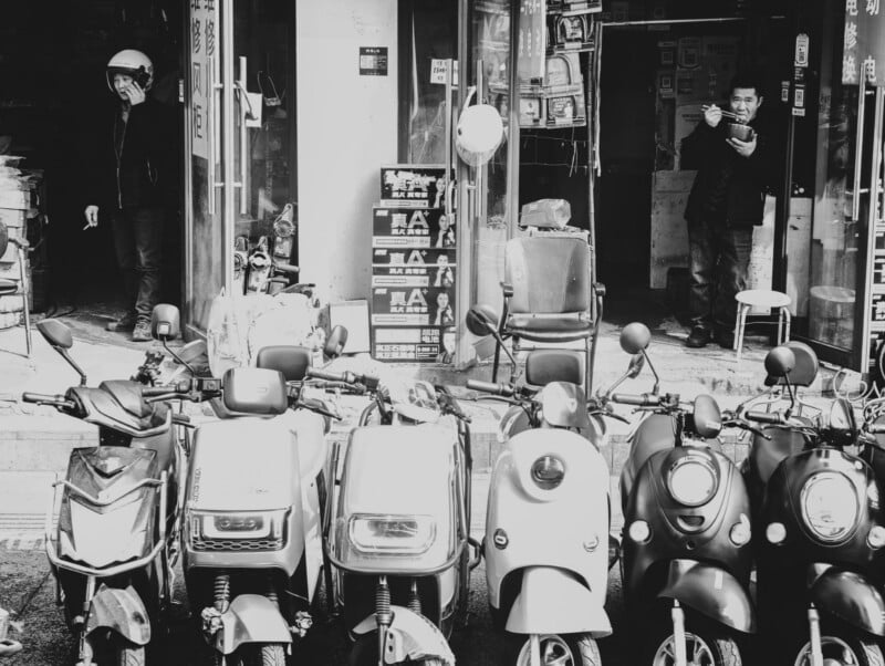 Black and white photo of scooters parked in front of shops; a man on the left wears a helmet and talks on his phone, while another man on the right stands in a doorway, appearing to adjust something near his face.
