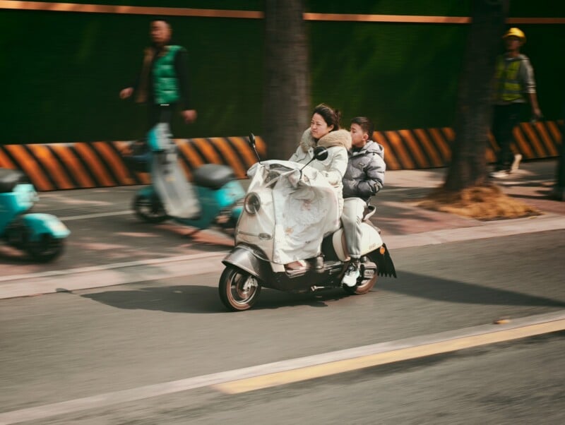A woman and a child ride a white scooter down a city street. The woman wears a thick blanket, and both appear bundled for cold weather. People and other scooters are seen blurred in the background, suggesting movement.
